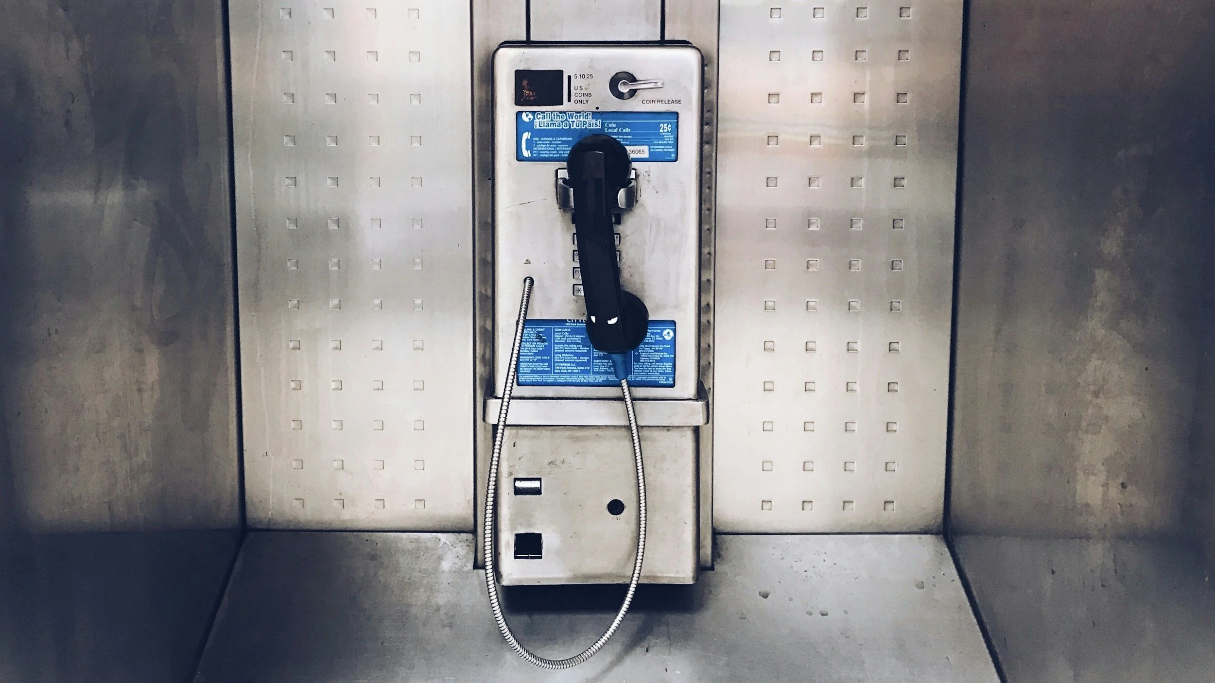 An old public telephone booth with a black rotary phone mounted on a metallic display with instructions and a coin slot, inside a metallic enclosure.