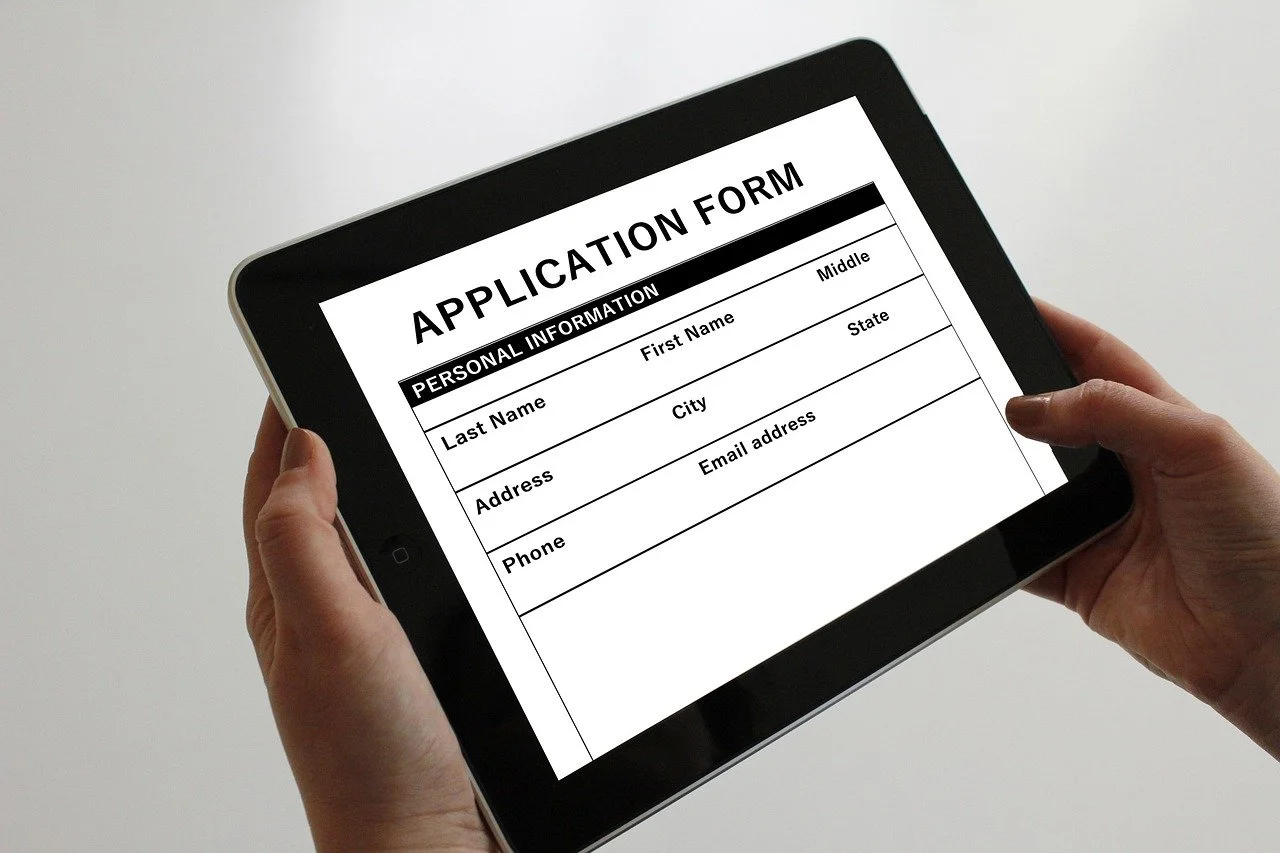 Hand holding tablet displaying an application form with sections for personal information such as last name, first name, middle name, address, city, state, phone, and email address.