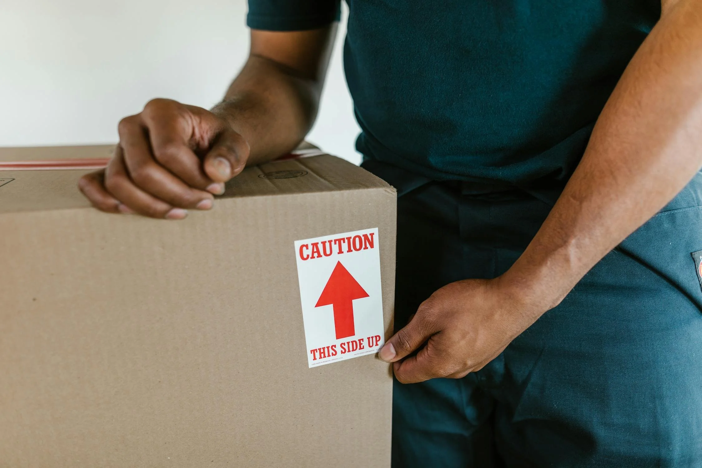 Person handling a cardboard box with a caution label that has an upward arrow and the words 'Caution This Side Up'.