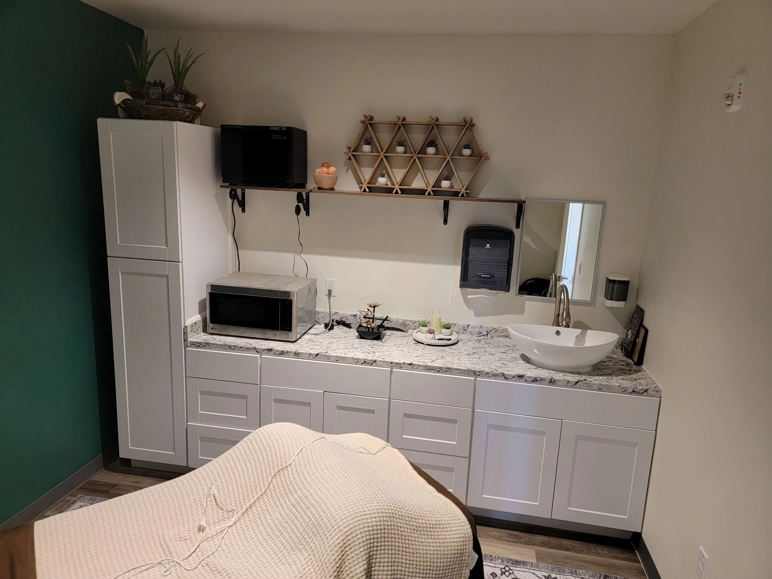 A well-organized spa treatment room featuring a countertop with a white sink, granite surface, cabinets, a microwave, and decorative items. There's a small triangular shelf with plants and a hand towel dispenser. A massage table with a white blanket
