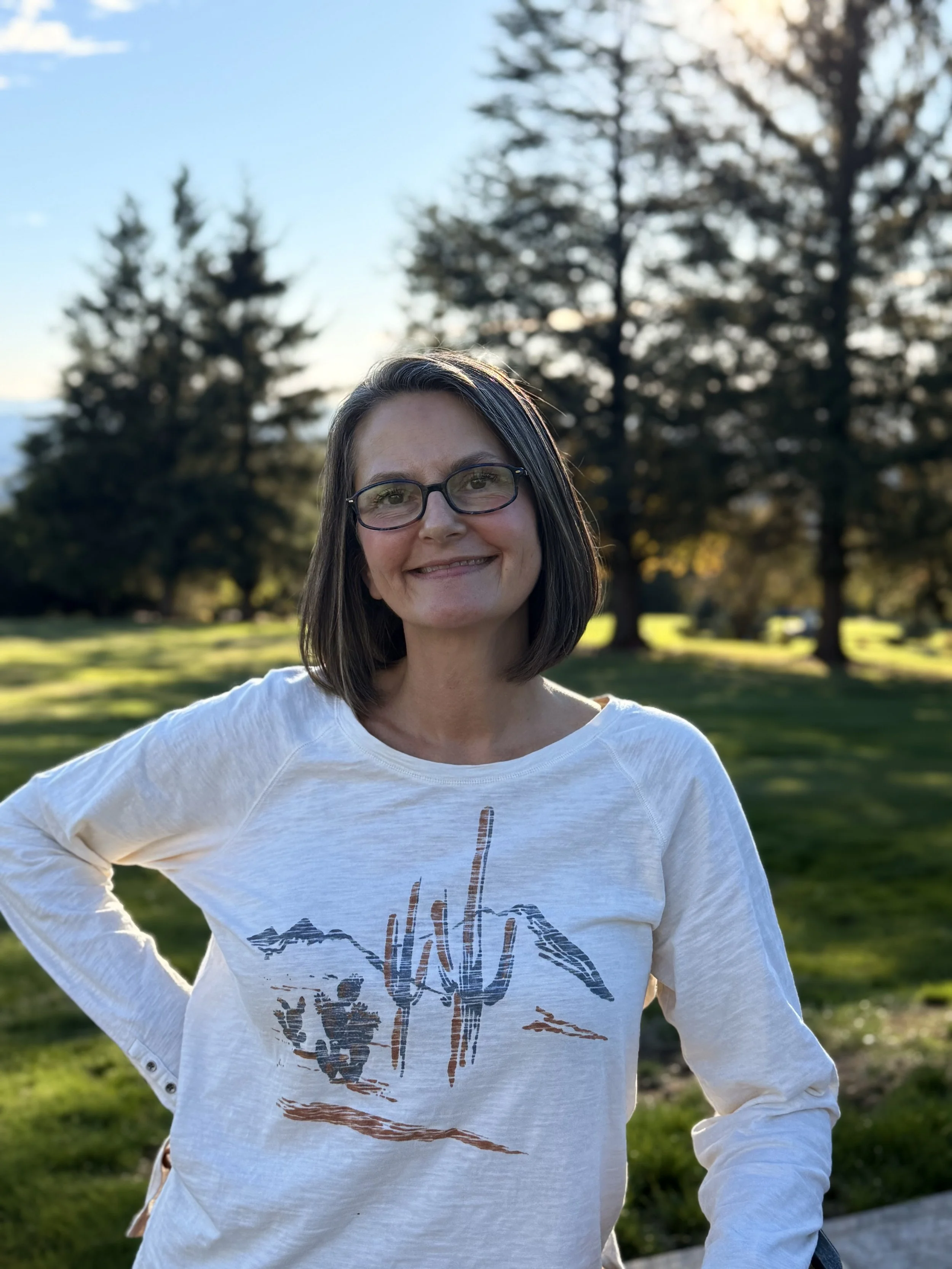 A woman with glasses and shoulder-length dark hair smiling outdoors in a park during sunset, with trees and grass in the background.