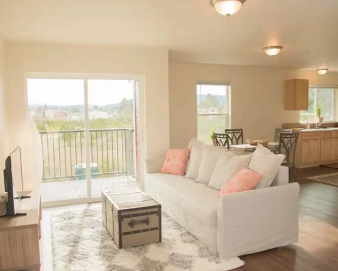 Bright living room with sliding glass door leading to a balcony, white sofa with pink and white pillows, wooden trunk coffee table, area rug, dining table and chairs, kitchen with wooden cabinets, and multiple ceiling lights.