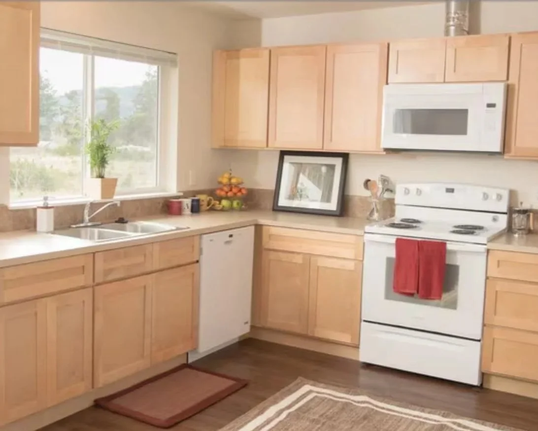 A kitchen with wooden cabinets, a white stove with a red towel hanging, a microwave above the stove, a white dishwasher, and a window with a potted plant. There are fruit bowls on the counter, and a framed picture leaning against the wall.