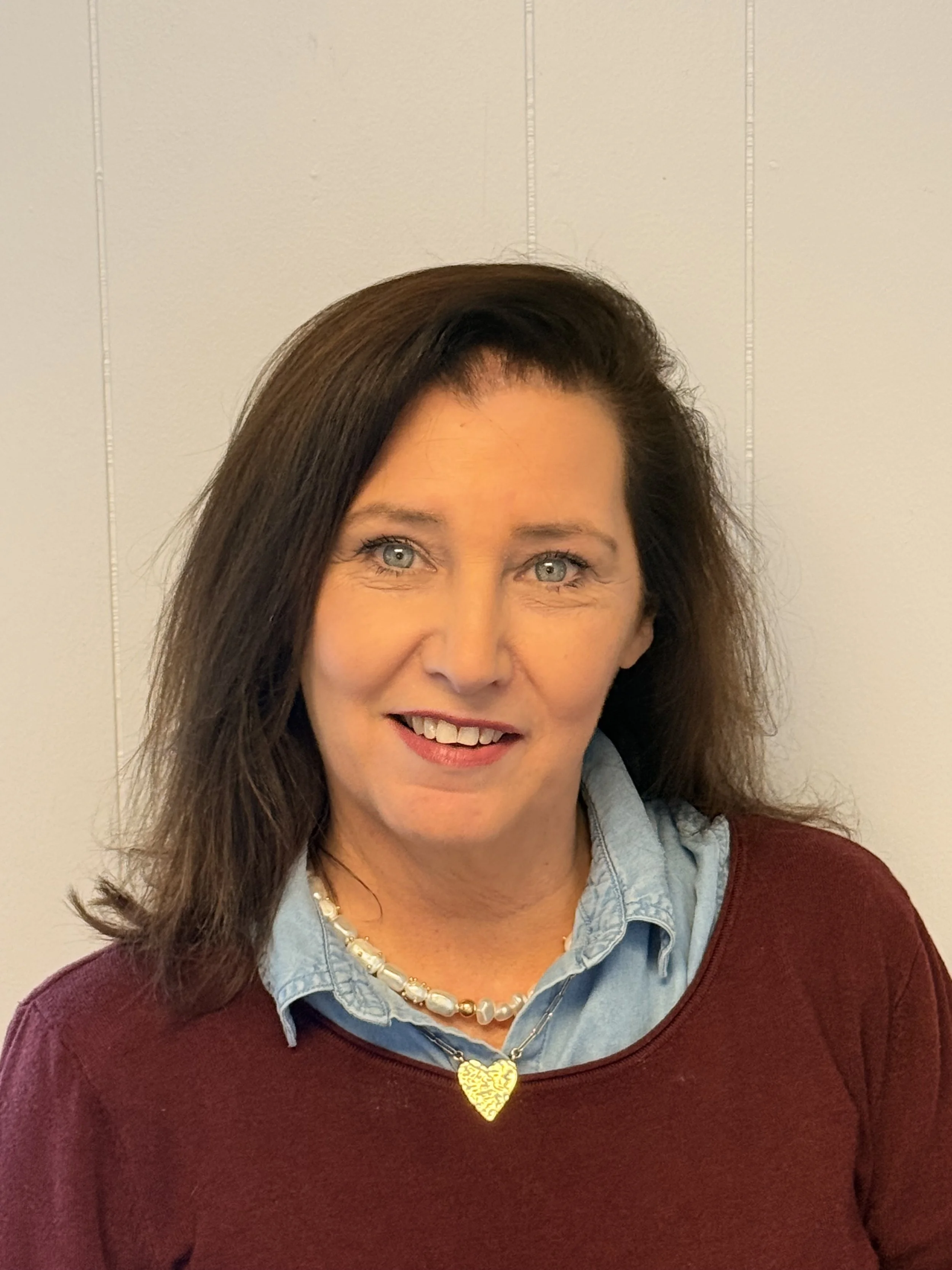 A woman with shoulder-length brown hair, blue eyes, wearing a light blue collared shirt under a maroon sweater, a pearl necklace with a gold heart pendant, smiling against a plain light-colored wall.