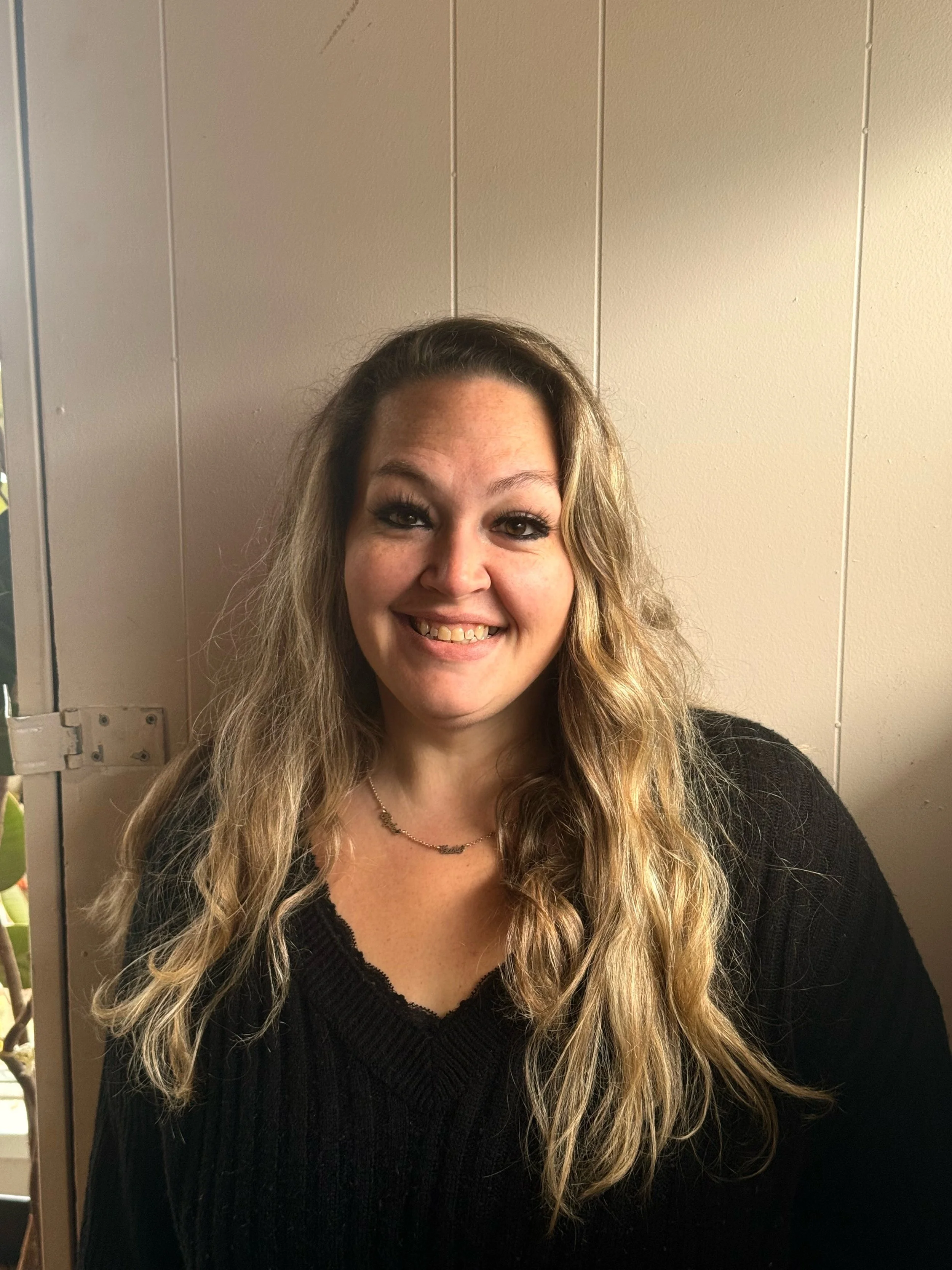 A woman with long, wavy blonde hair smiling at the camera, wearing a black sweater and a delicate necklace, standing against a plain beige wall.