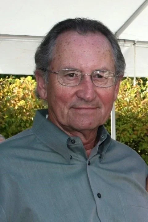A middle-aged man with glasses and gray hair, wearing a button-up shirt, standing outdoors under a white tent with greenery in the background.