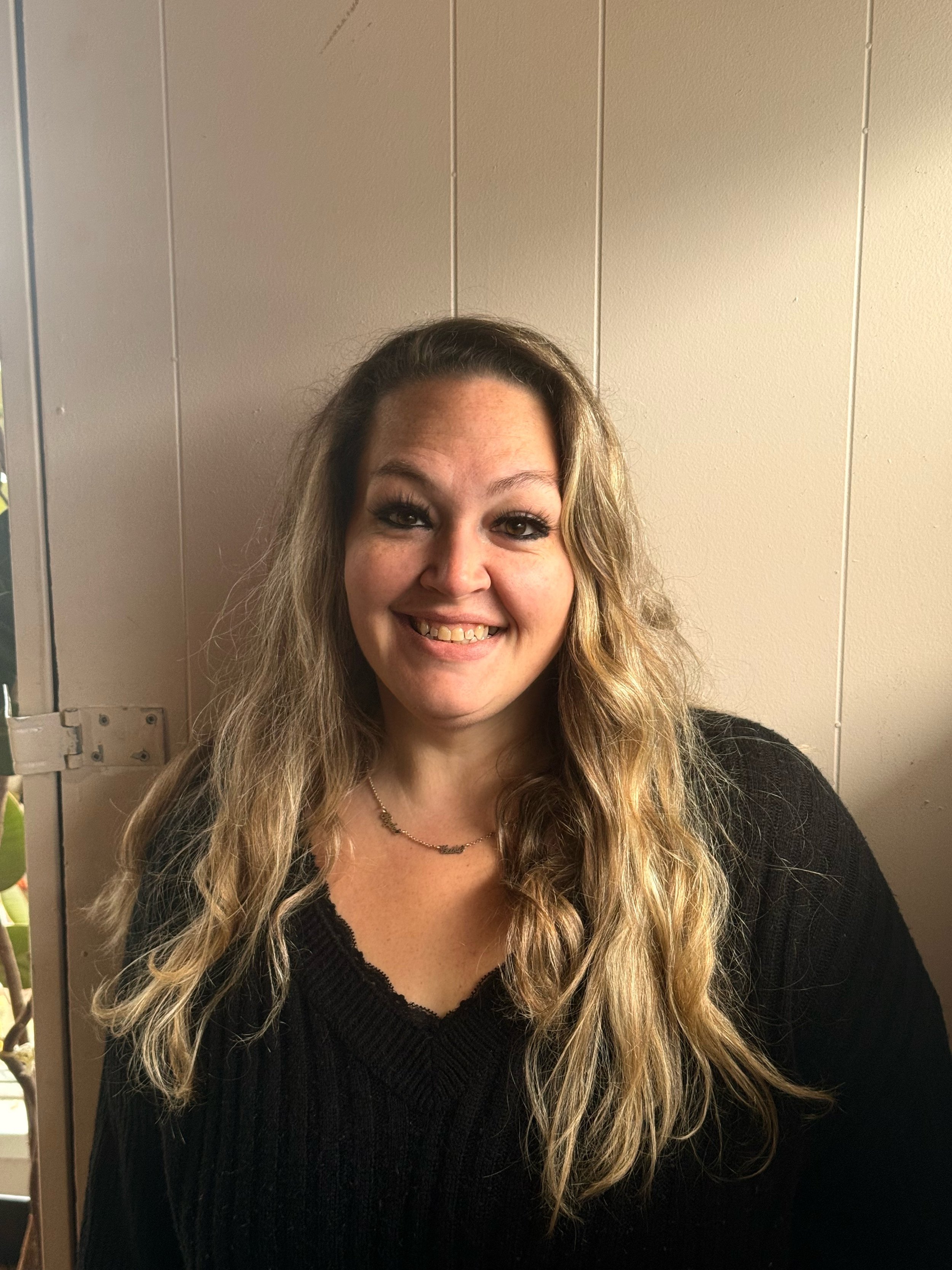 Portrait of a smiling woman with long wavy blonde hair, wearing a black sweater and a necklace, standing against a beige wall.