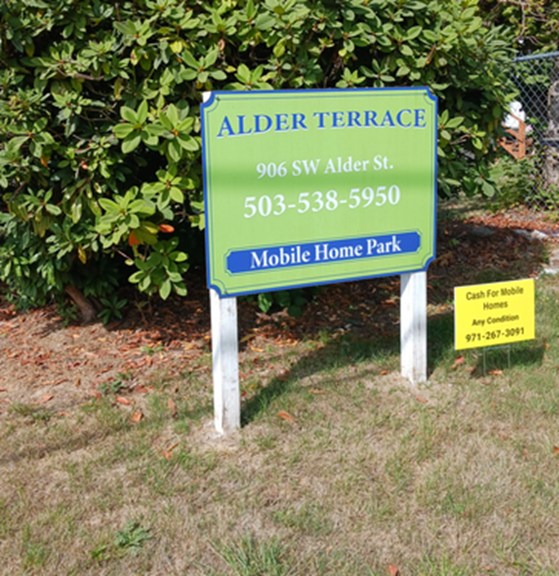 Sign for Alder Terrace Mobile Home Park with address and phone number, located near bushes and grass.