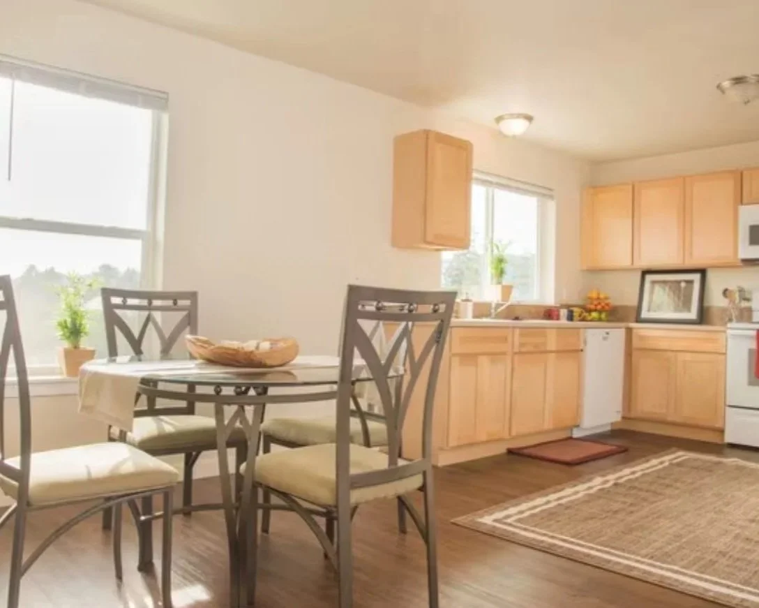Kitchen and dining area with a round table, four chairs, light wood cabinets, window with a plant, and a rug on the hardwood floor.