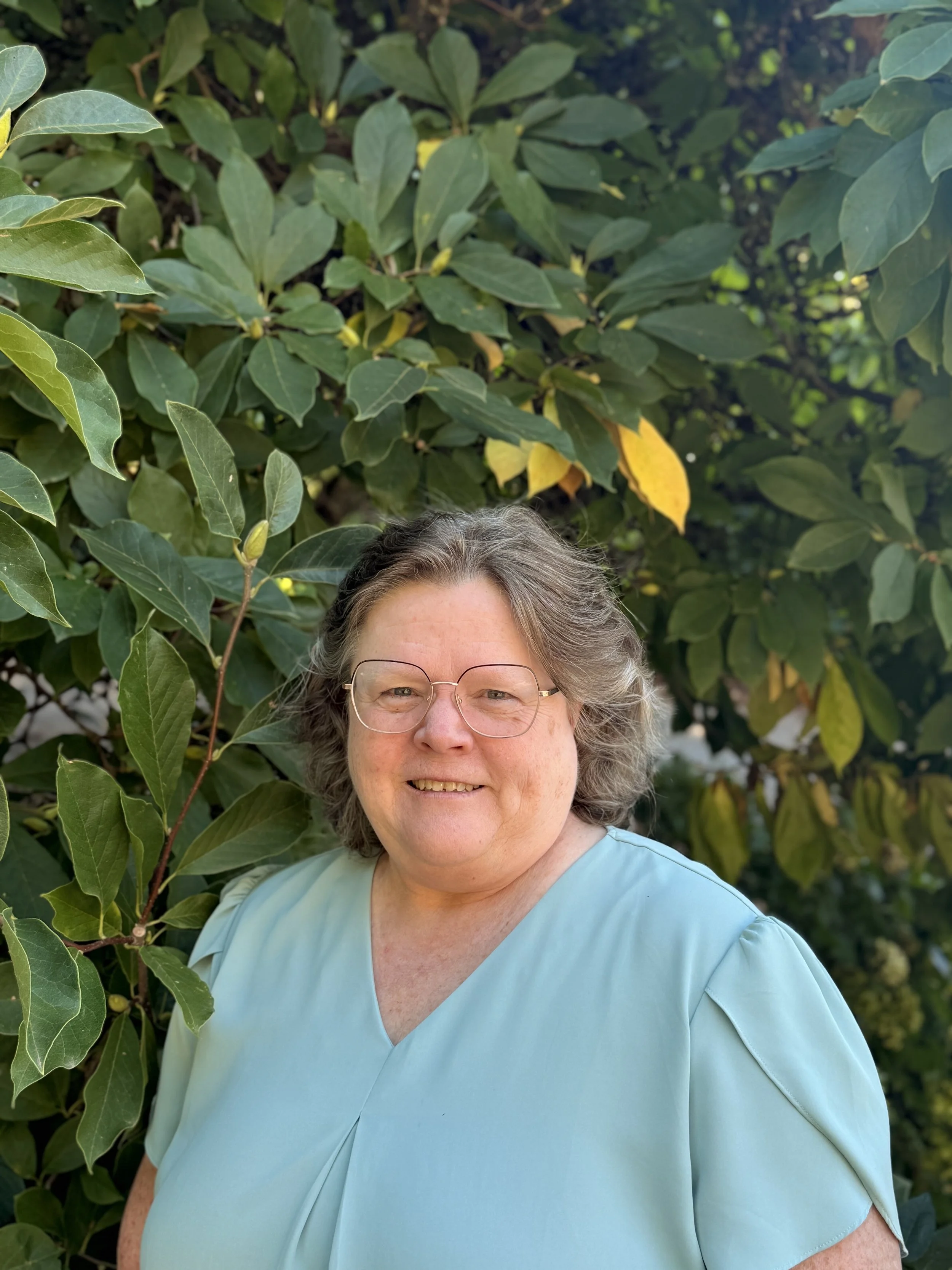 A smiling woman with glasses and shoulder-length gray hair standing outdoors in front of green leafy bushes.