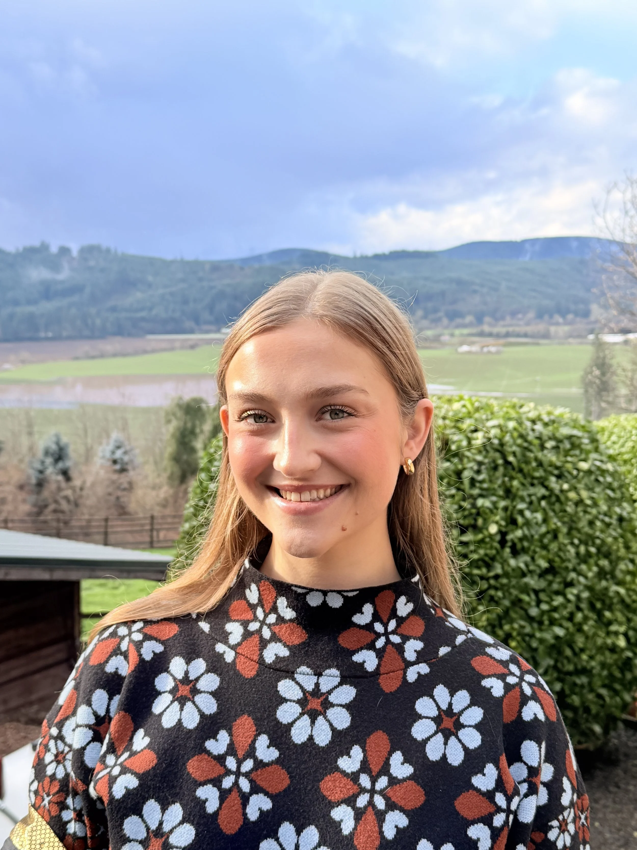 A man with dark, curly hair, glasses, and a beard, wearing a dark green sweater, sits indoors with a neutral expression, near a white wall and a large green plant in the background.