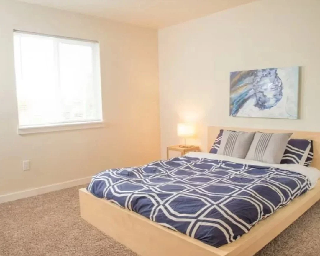 Bedroom with a bed featuring a blue and white geometric patterned comforter, three striped pillows, a wooden nightstand with a lamp, and abstract artwork on the wall, with a window on the left side.