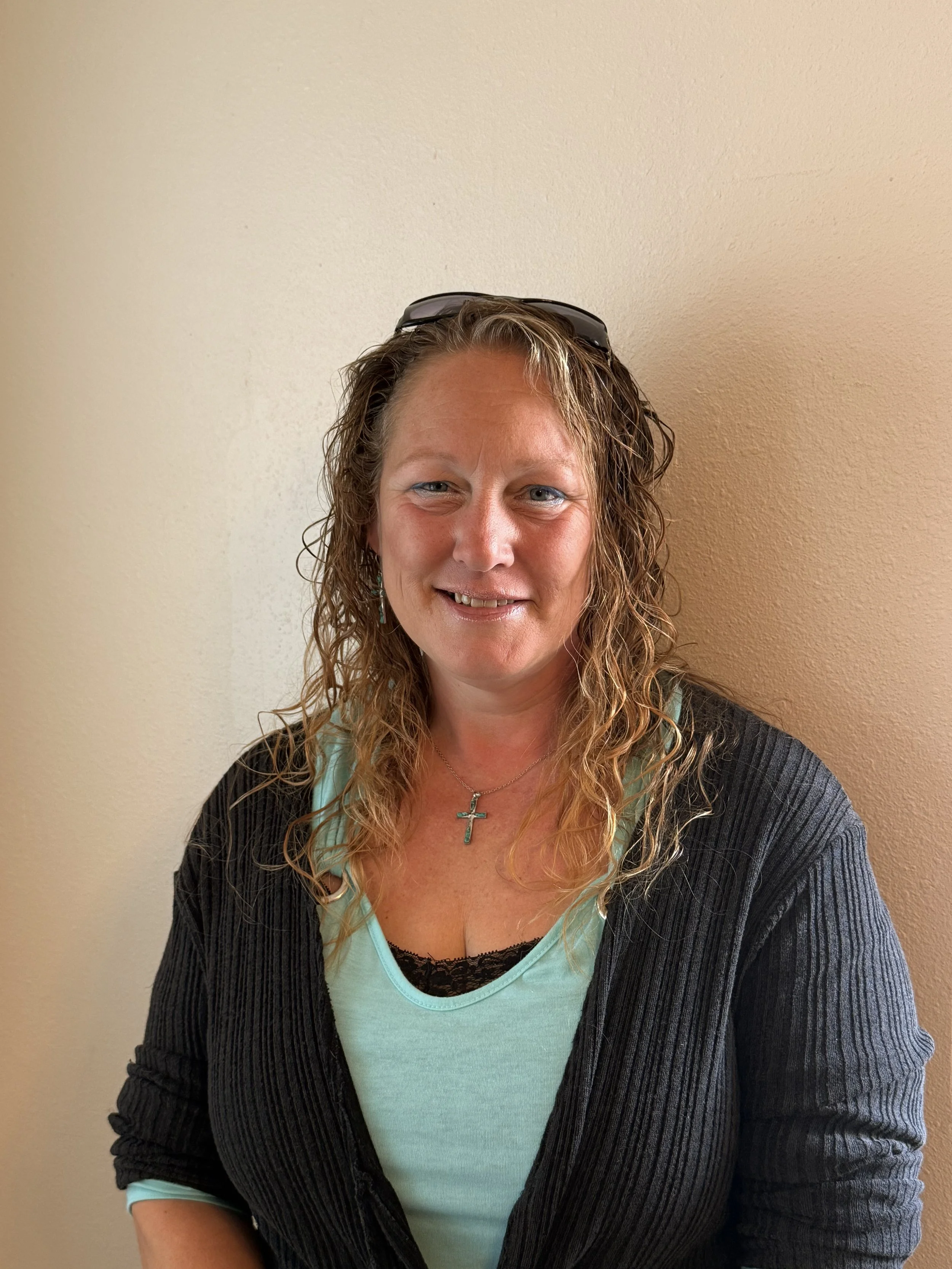 A woman with curly, shoulder-length hair wearing glasses on her head, a turquoise shirt with black lace trim, a black cardigan, and a cross necklace, standing against a beige wall, smiling at the camera.