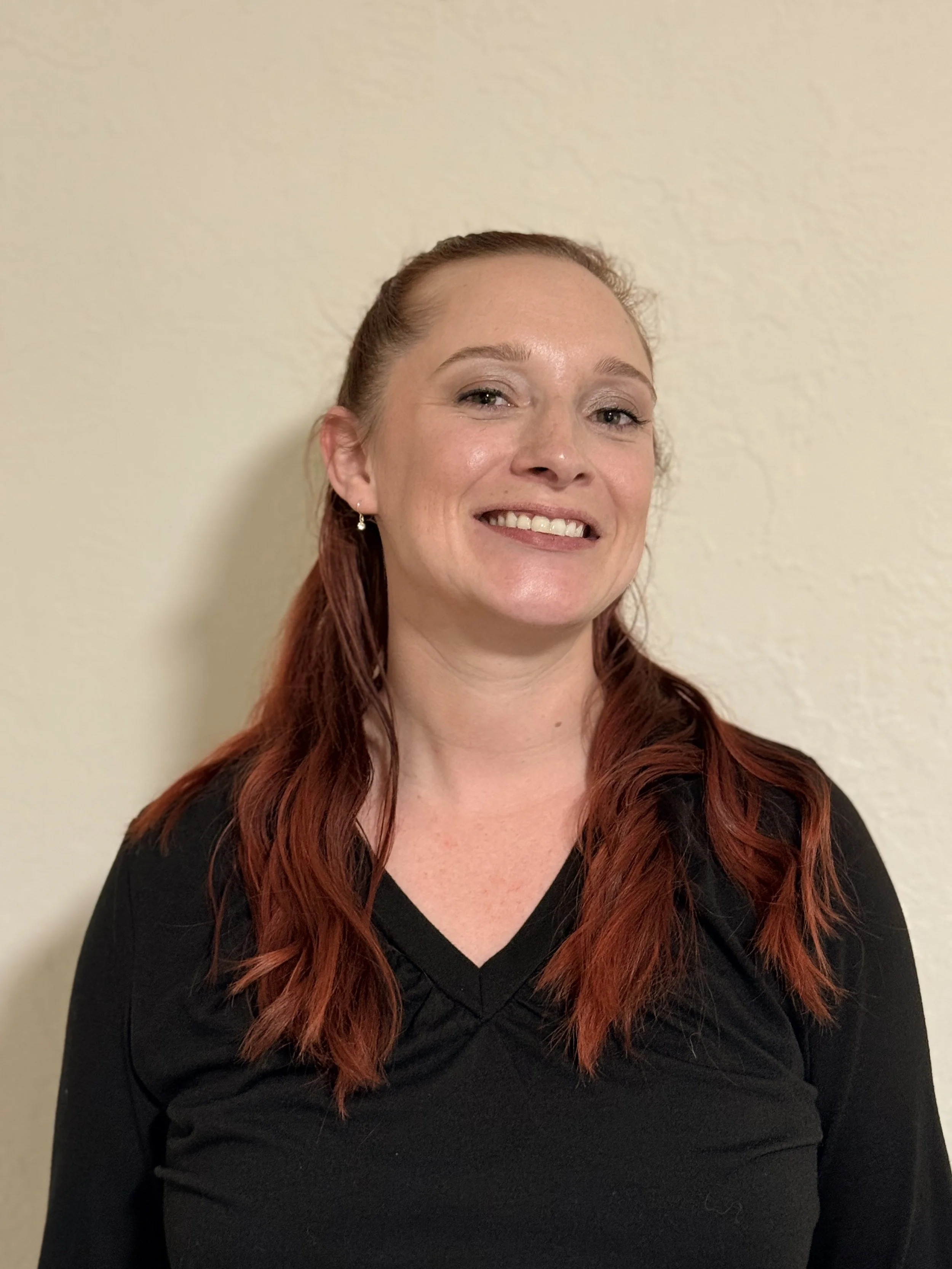A woman with red hair smiling, wearing a black top, standing against a beige wall.
