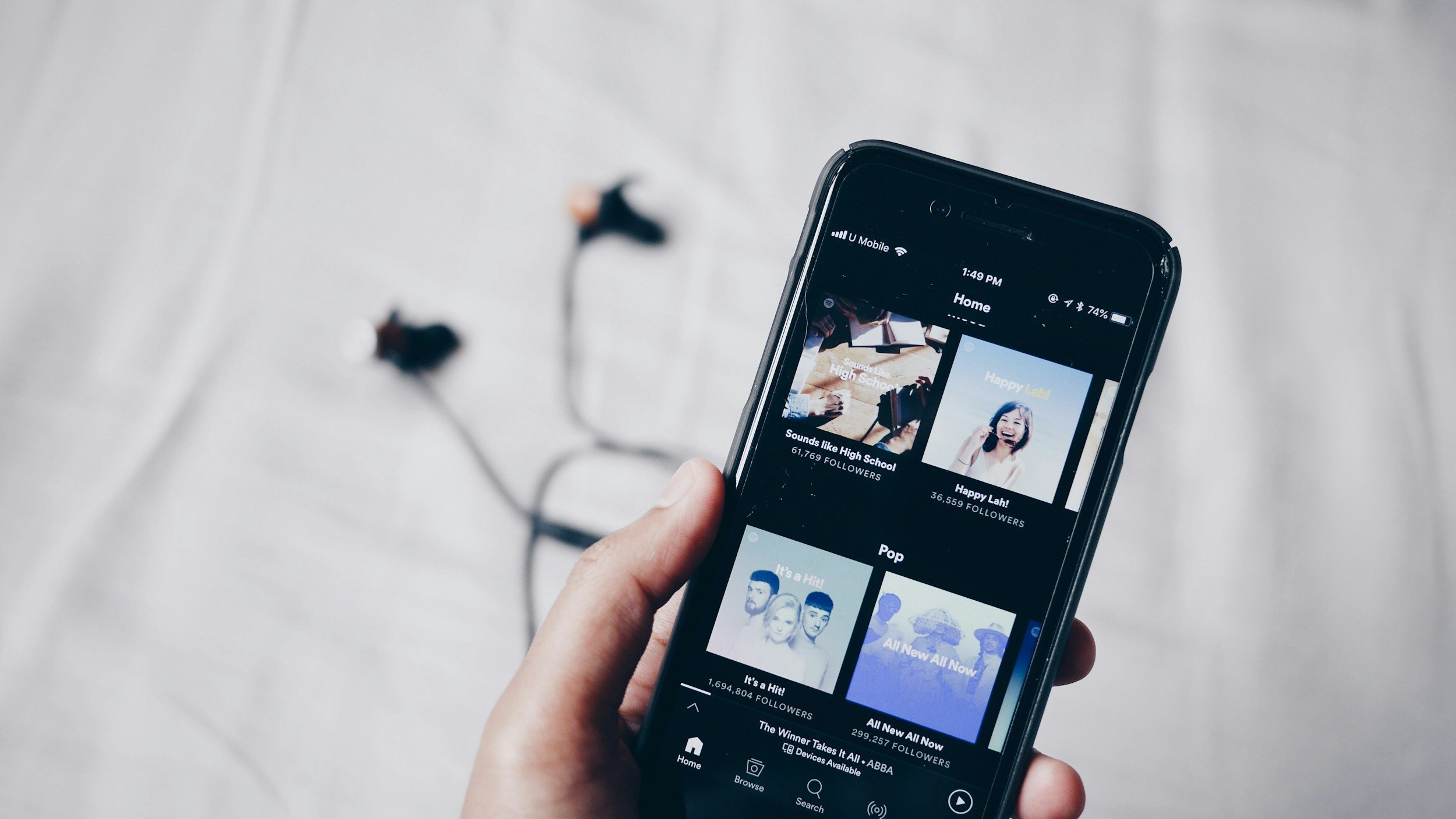 Main tenant un smartphone affichant une application de musique avec des écouteurs en arrière-plan flou.