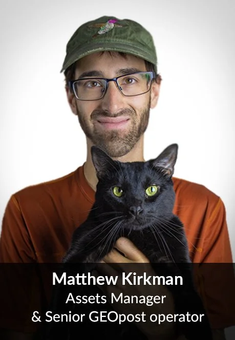 A man wearing glasses and a green cap with a bird embroidered on it, holding a black cat with green eyes, against a plain gray background.