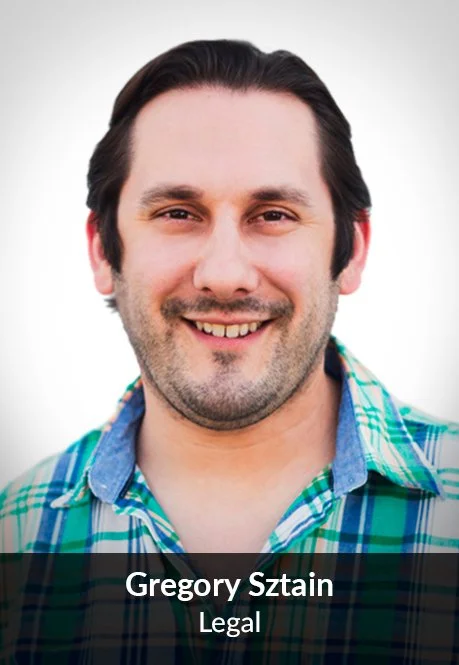 A man with dark hair and facial hair, smiling, wearing a blue, green, and white plaid shirt against a plain light background.