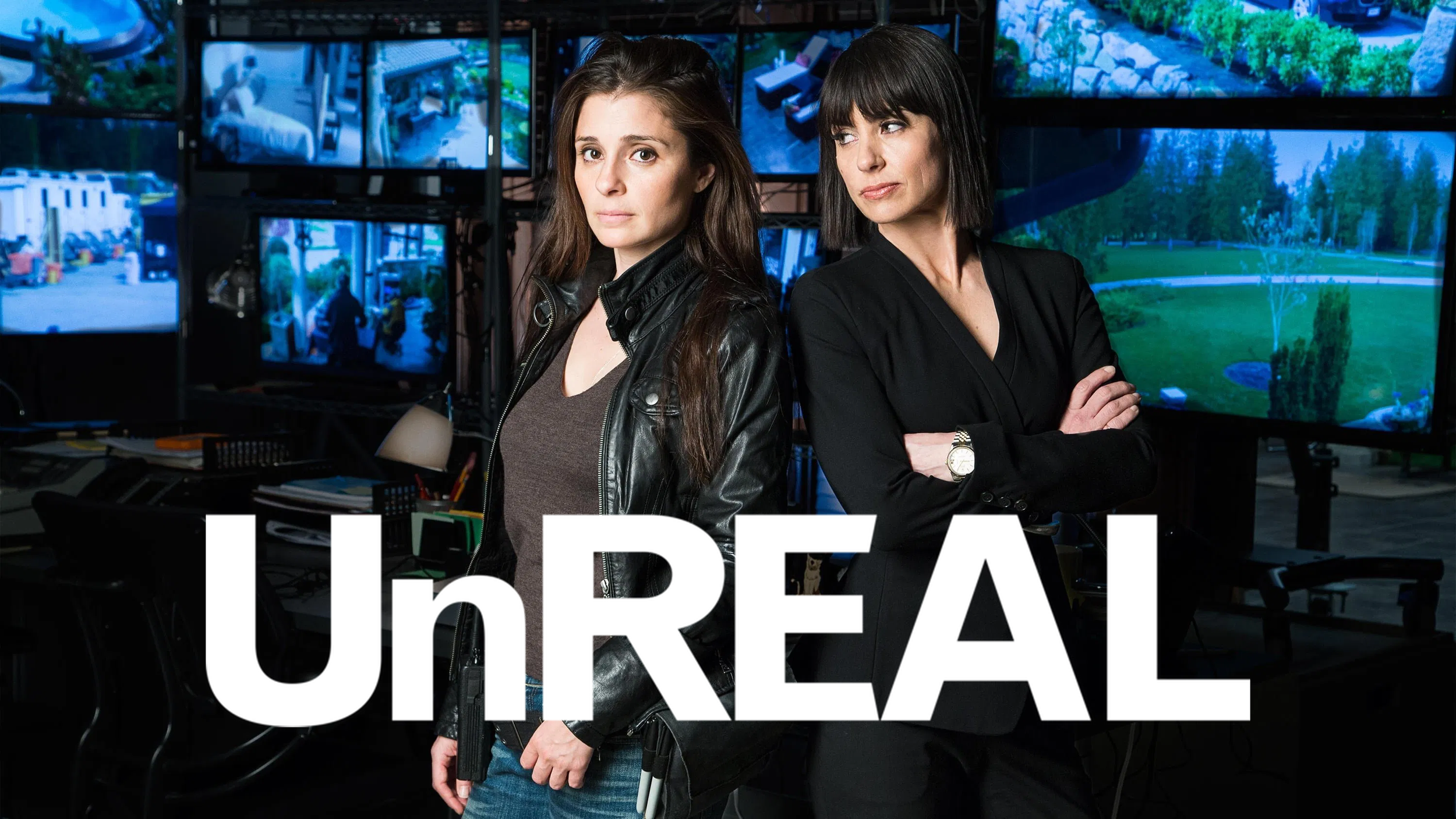 Two women stand back-to-back in a control room with multiple monitors displaying outdoor scenery, one wearing a leather jacket and the other in a black blazer, with the word 'UnREAL' prominently overlaid.