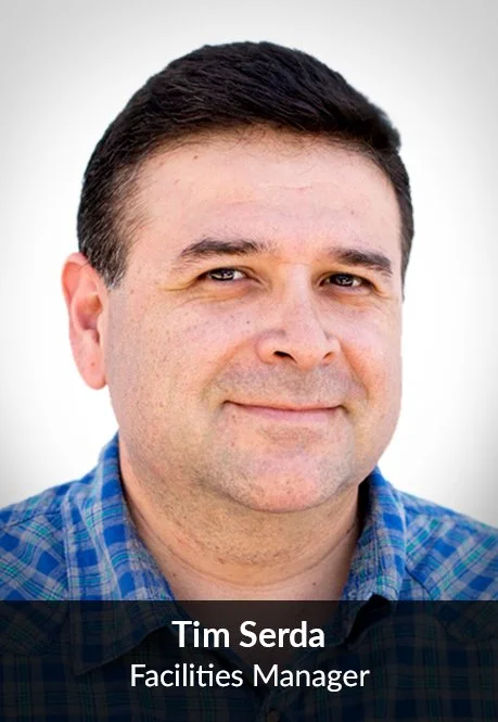 Headshot of a man with short dark hair, wearing a blue plaid shirt, smiling slightly, against a white background. Caption reads: 'Tim Serda, Facilities Manager.'