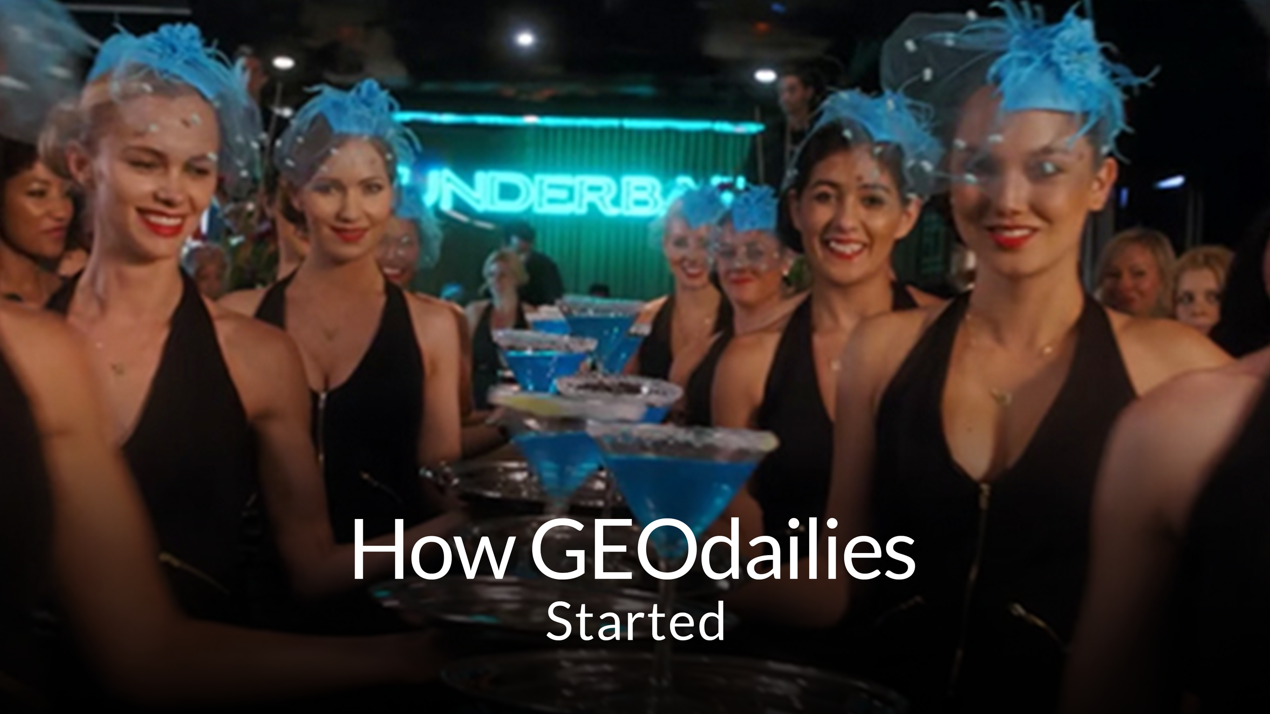 Women in black dresses with blue feathered headpieces smiling with drinks at a bar or club, neon sign in background reading 'UNDERBAR'.