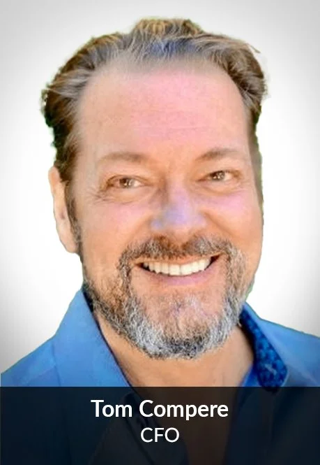 A headshot of Tom Compere, a man with brown hair and a beard, smiling and wearing a blue shirt.
