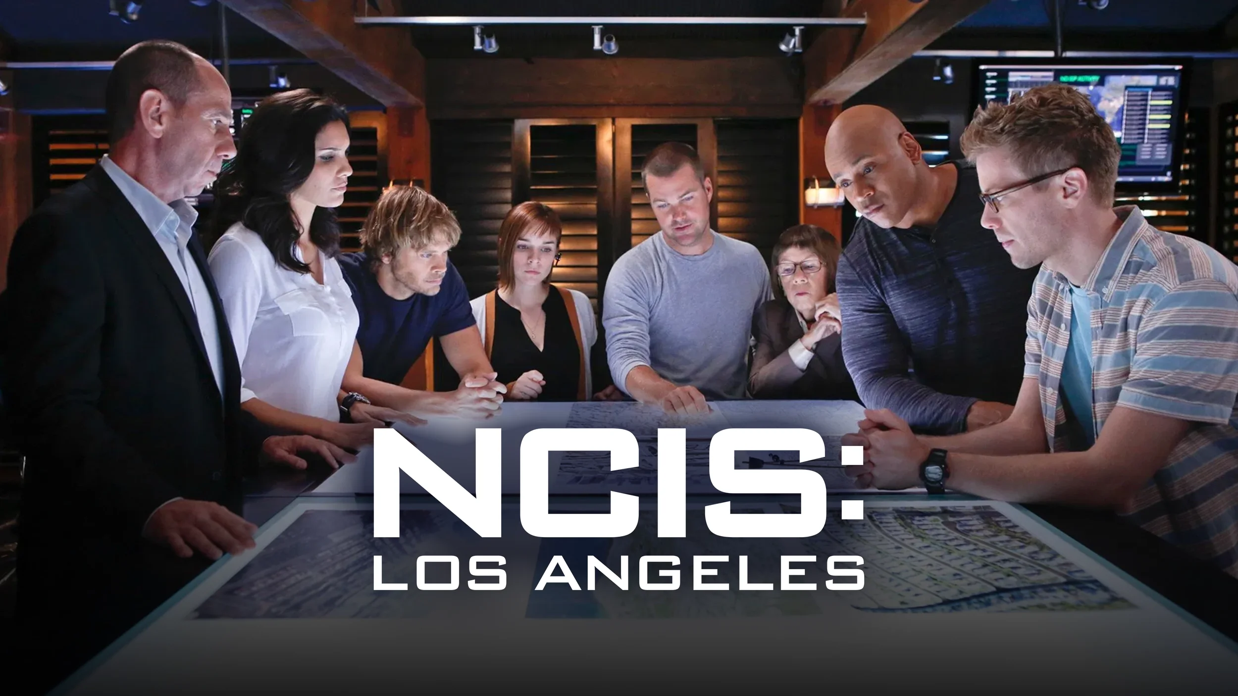 A group of people gathered around a table with documents, in a dark room with multiple screens in the background, discussing and analyzing information. The overlay text reads "NCIS: Los Angeles."