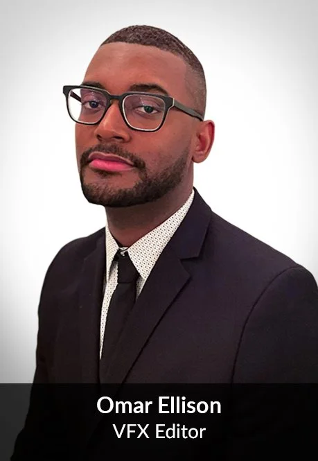 A man with short hair, wearing glasses, a dark suit, white shirt, and a tie, posing against a plain background.
