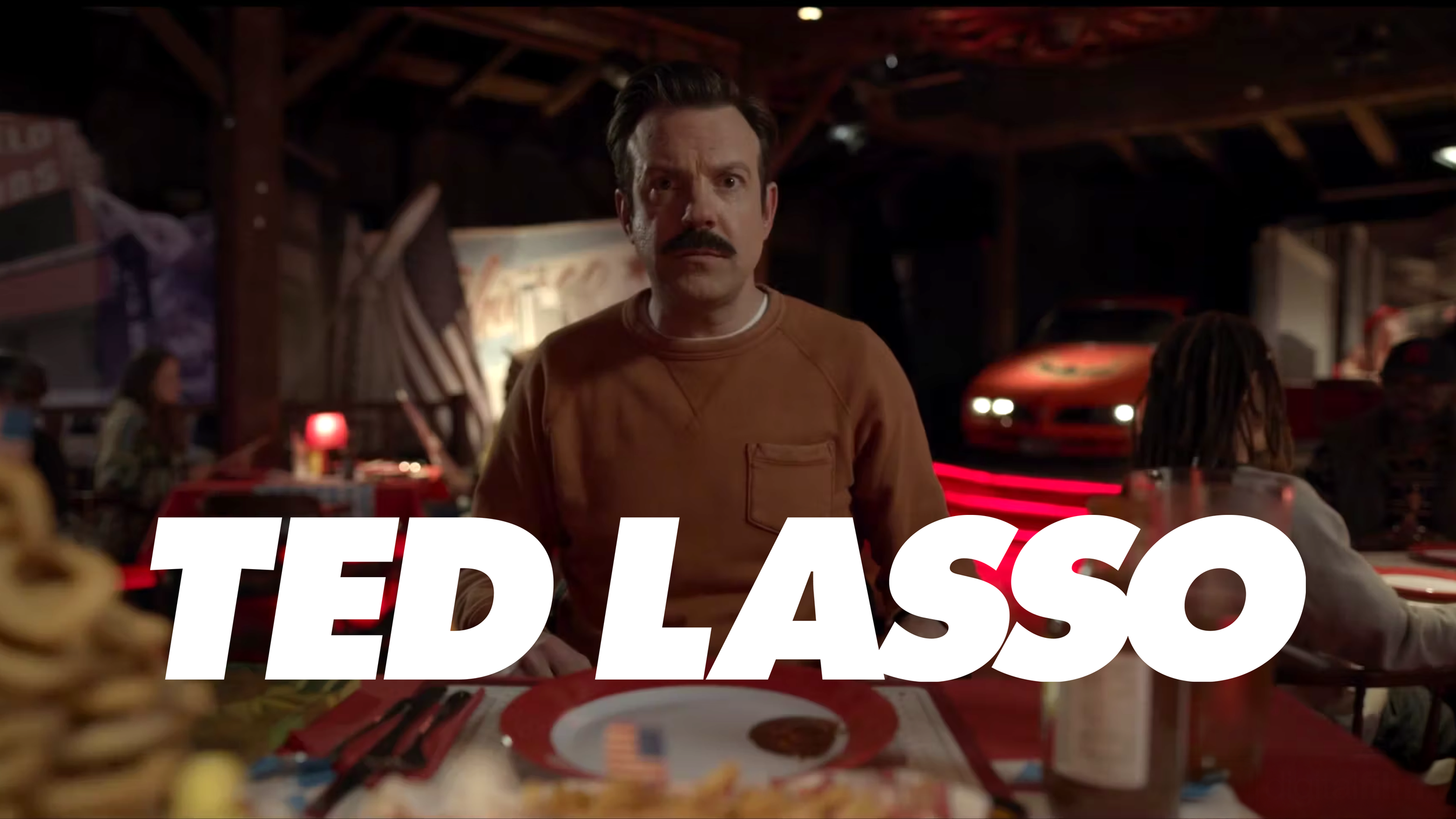 Still from the movie 'Ted Lasso' showing a man with a mustache sitting at a decorated table in a dimly lit restaurant or bar, with the text 'TED LASSO' overlaid.