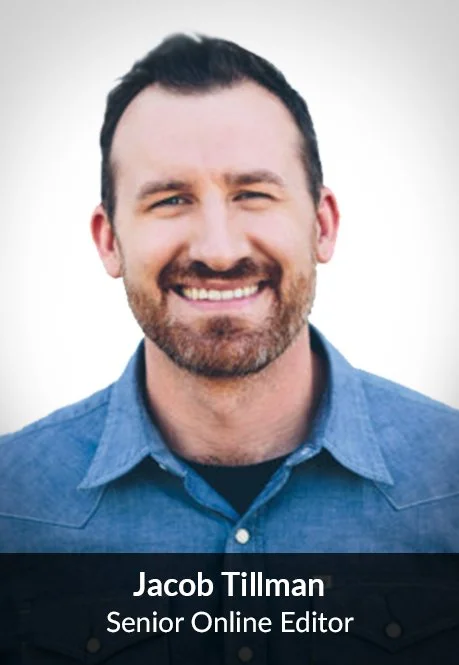 A man with dark hair and a beard smiling, wearing a blue button-up shirt, against a plain white background.