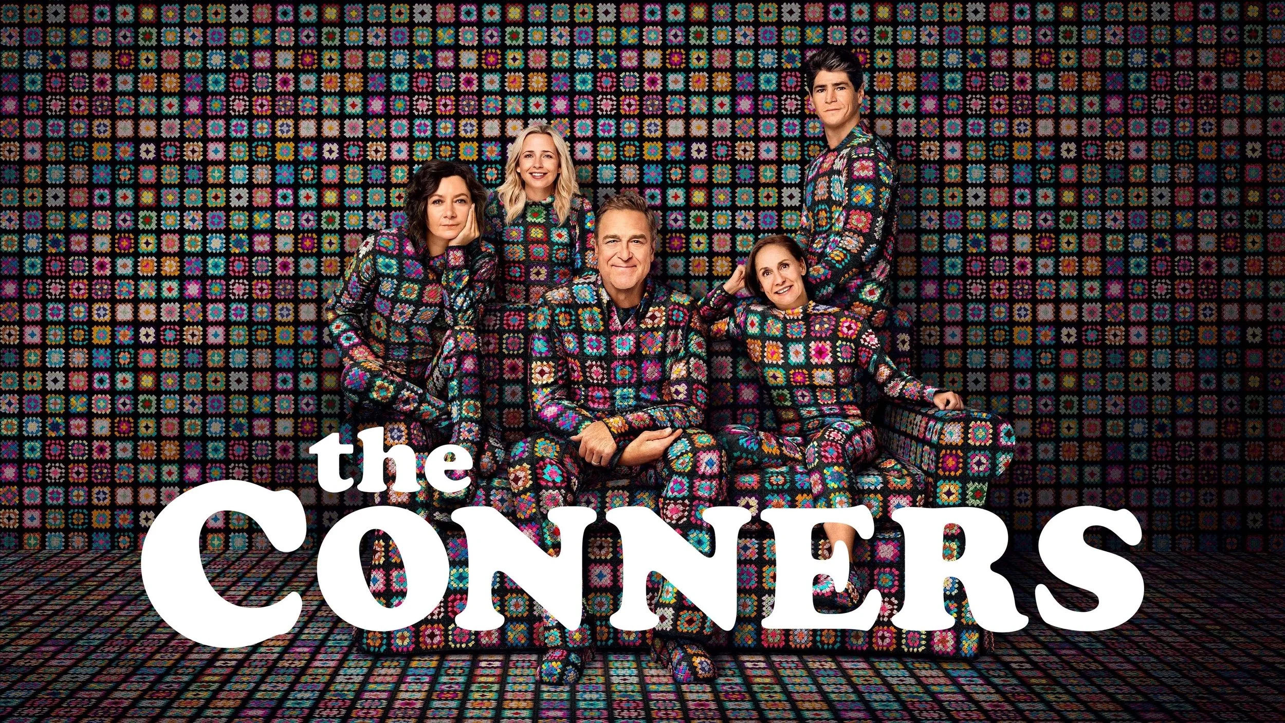 The main cast of the TV show 'The Conners' sitting and standing in front of a colorful quilted background, all wearing matching patterned outfits, with the show's title written in white text.
