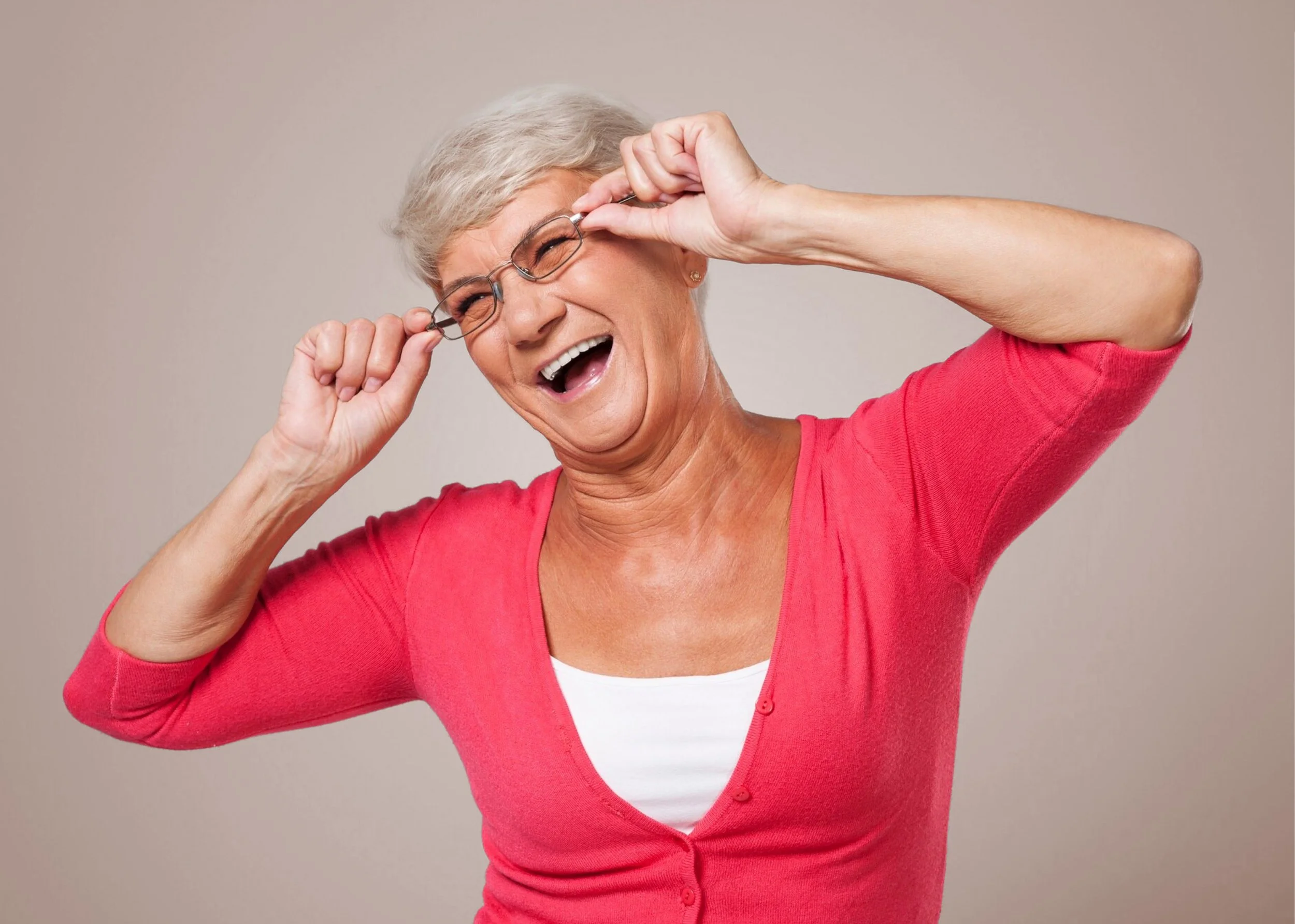 An elderly woman with short gray hair wears glasses and a pink cardigan, smiling and holding her glasses with both hands against a plain background.