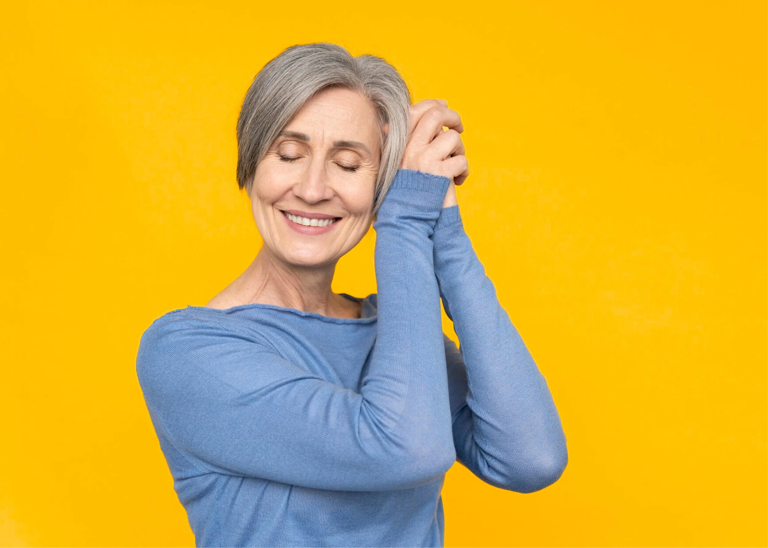 A smiling older woman with grey hair touching her head against a yellow background.