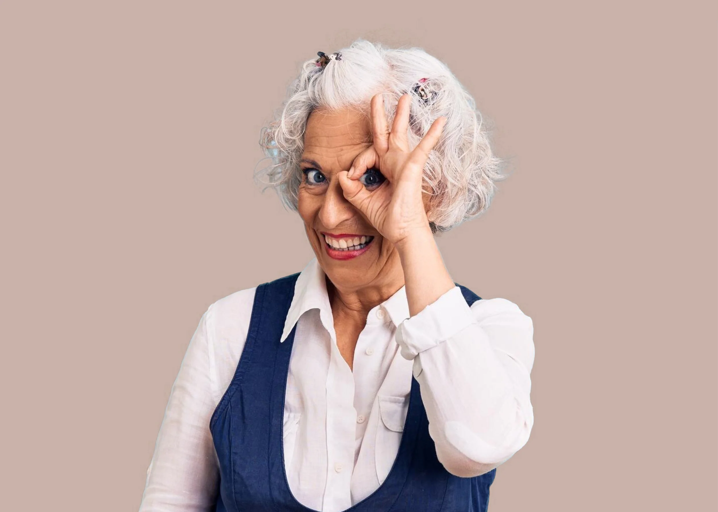 Older woman with curly gray hair making a playful gesture with her hand around her eye, smiling, wearing a white shirt and a dark vest, standing against a plain beige background.