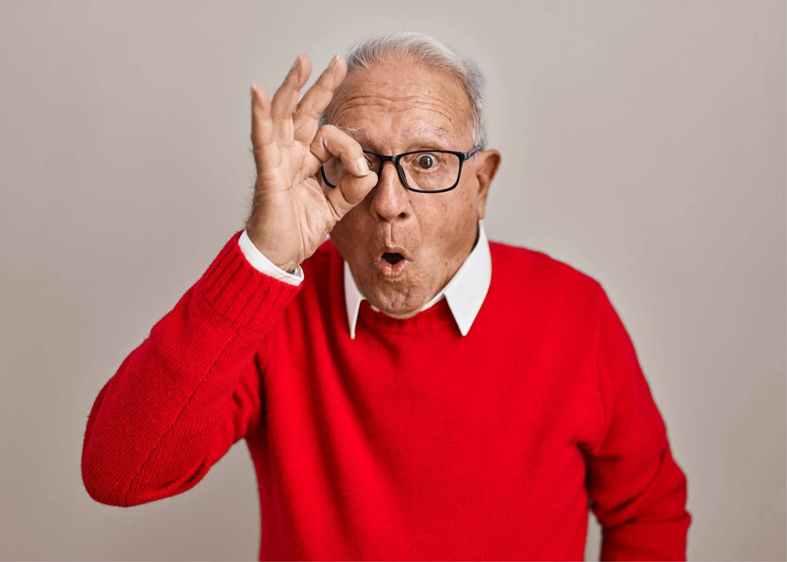 An elderly man wearing a red sweater and glasses making an 'OK' gesture around his eye with a surprised expression.