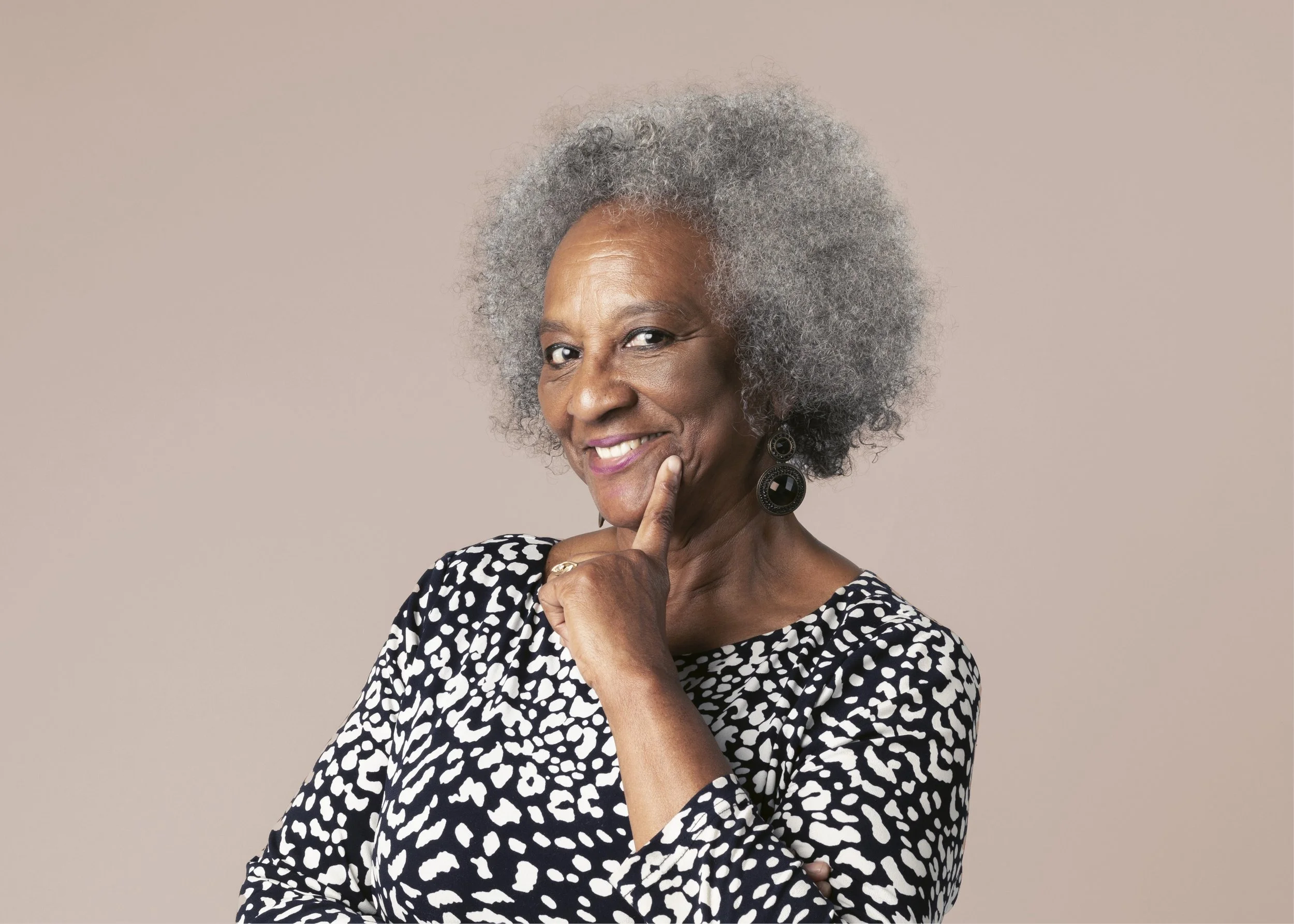 Portrait of an elderly woman with gray afro hair, wearing a black and white patterned top, smiling and touching her chin with one finger, against a light beige background.