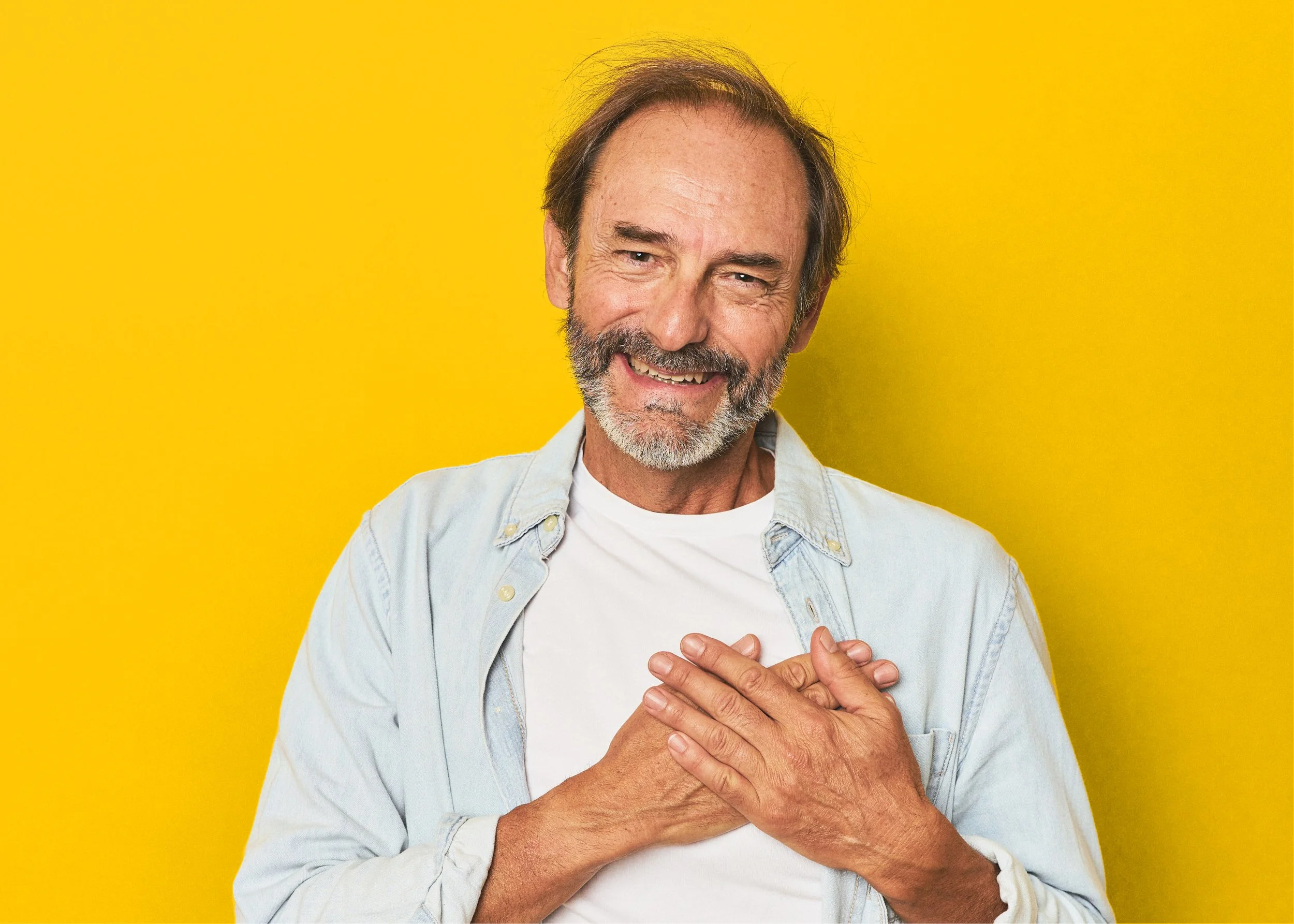 Smiling elderly man with a beard and mustache, wearing a light denim shirt over a white t-shirt, holding his hands over his chest against a bright yellow background.