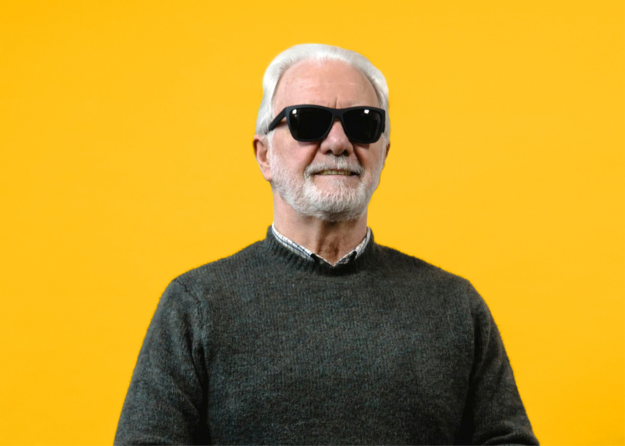 Older man with white hair and beard wearing black sunglasses, a dark sweater, and a collared shirt against a yellow background.