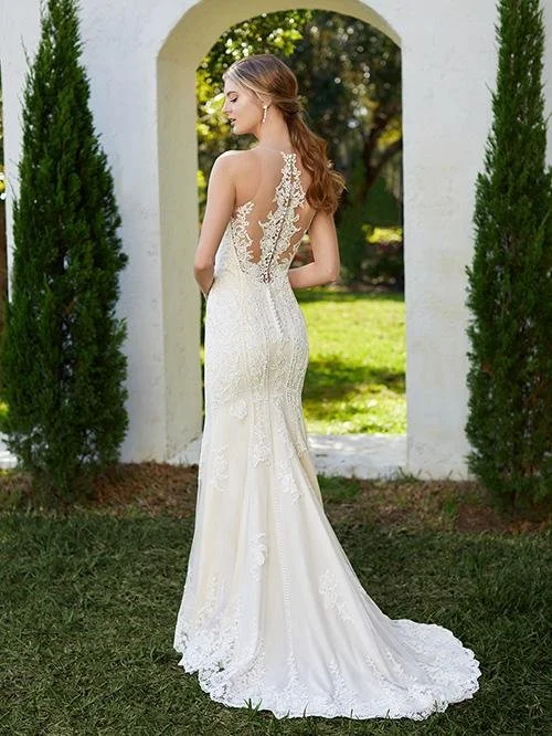 A woman in a white lace wedding dress standing outdoors under an arched doorway, with green trees and grass in the background.