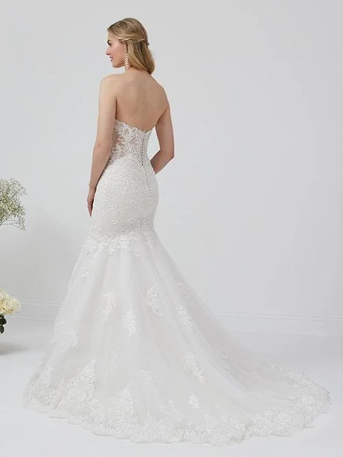 A woman in a white strapless wedding dress with lace details, standing in a plain light-colored room, facing away from the camera.