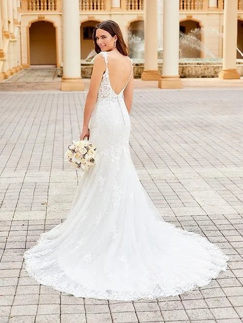 Brunette bride in a white lace wedding gown standing outdoors on a stone-paved courtyard, holding a bouquet of white and purple flowers, with a grand building featuring columns in the background.