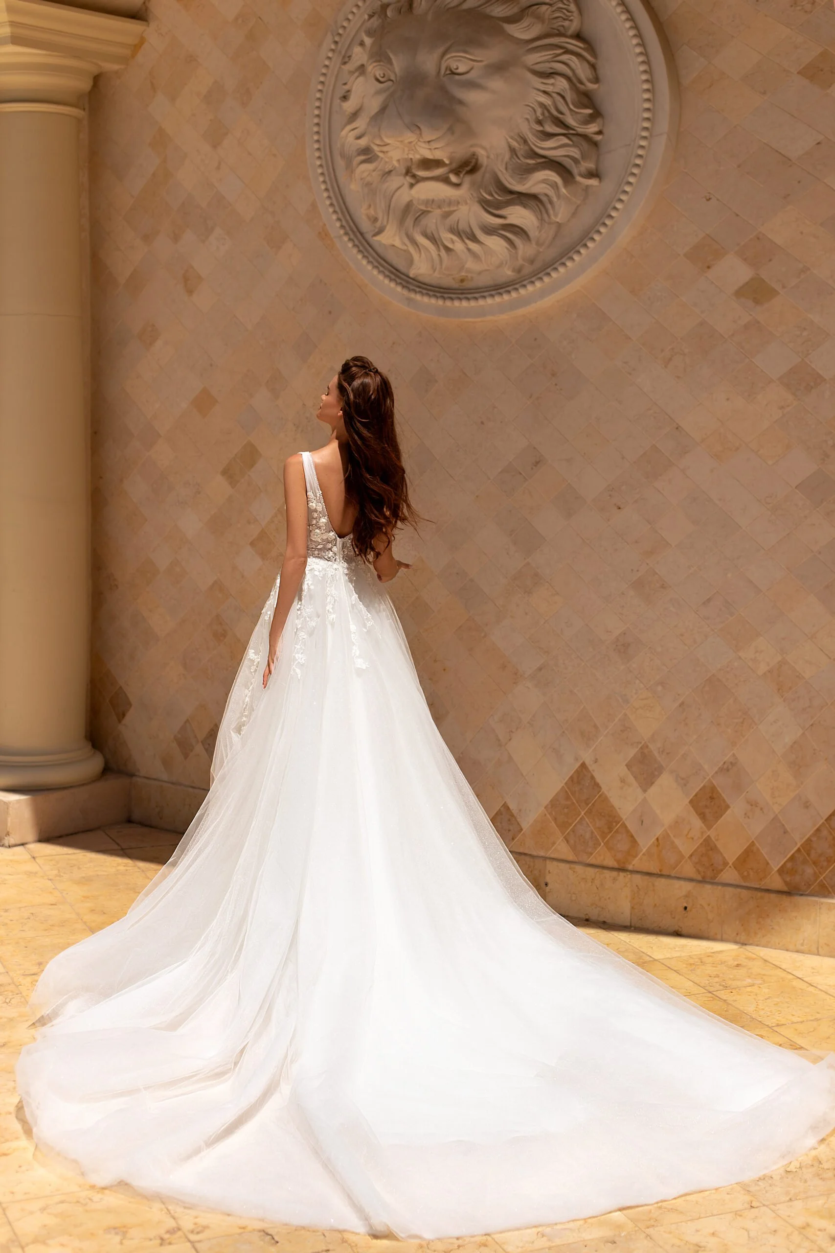 A woman in a white wedding dress with a long train standing indoors, facing left, in front of a beige tiled wall with a large circular sculpture of a lion's head.