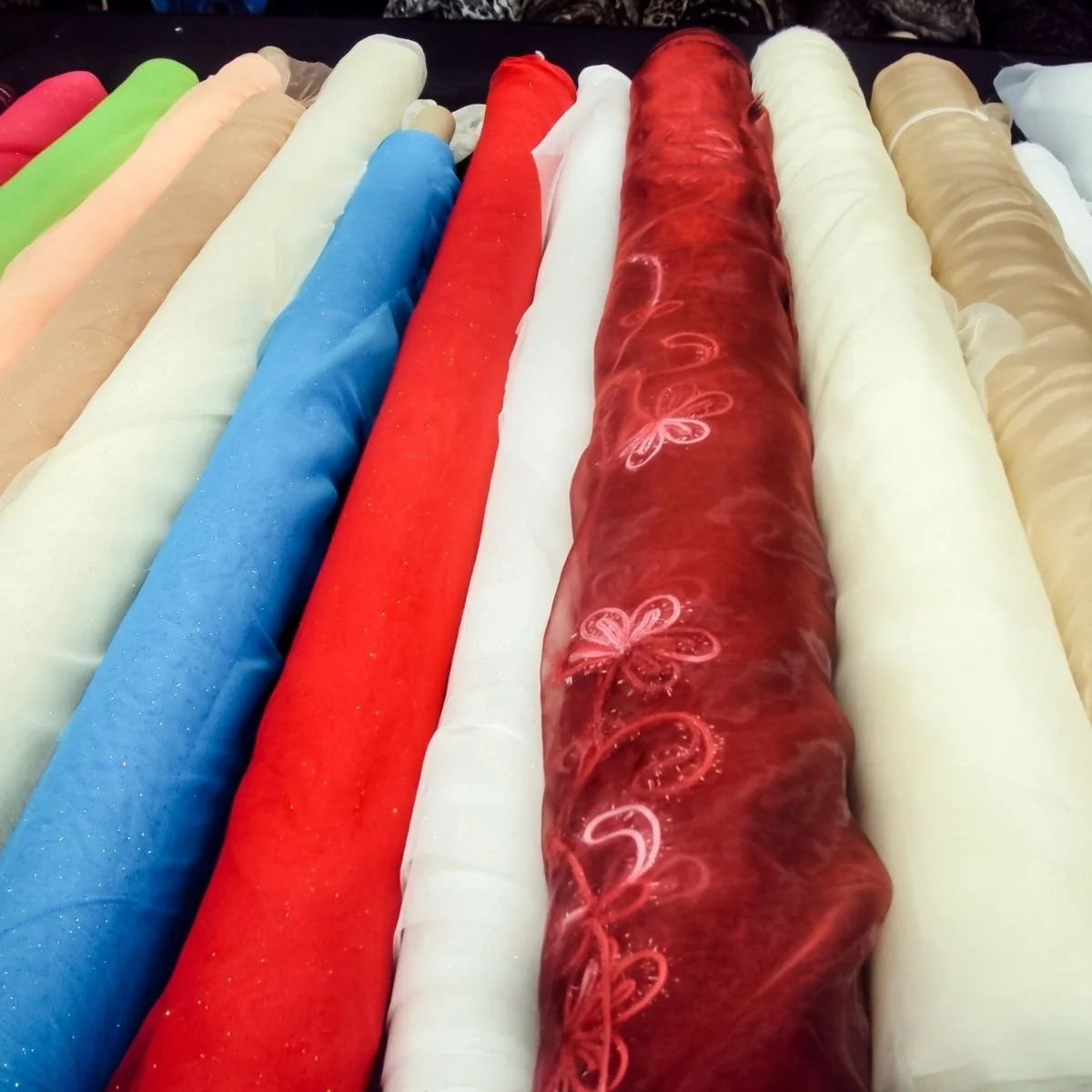 Multiple rolls of fabric in various colors, including green, beige, blue, red, white, and beige with floral embroidery, arranged side by side.