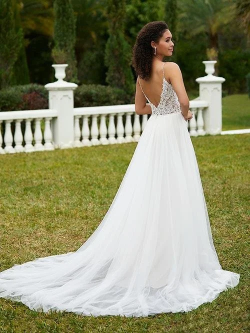 A woman in a white wedding dress with a lace bodice and flowing skirt, standing outside near a white railing and lush green trees.