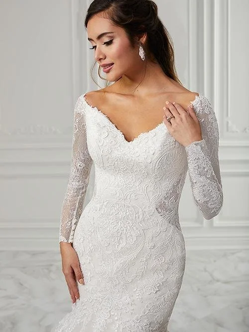 A woman in a white lace wedding dress with long sleeves and a V neckline, standing in a room with white walls, looking down with a gentle smile, wearing earrings and a ring.