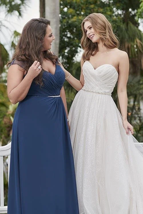 Two women in formal dresses smiling and looking at each other outdoors, one in a navy blue gown and the other in a white wedding dress with a beaded waist