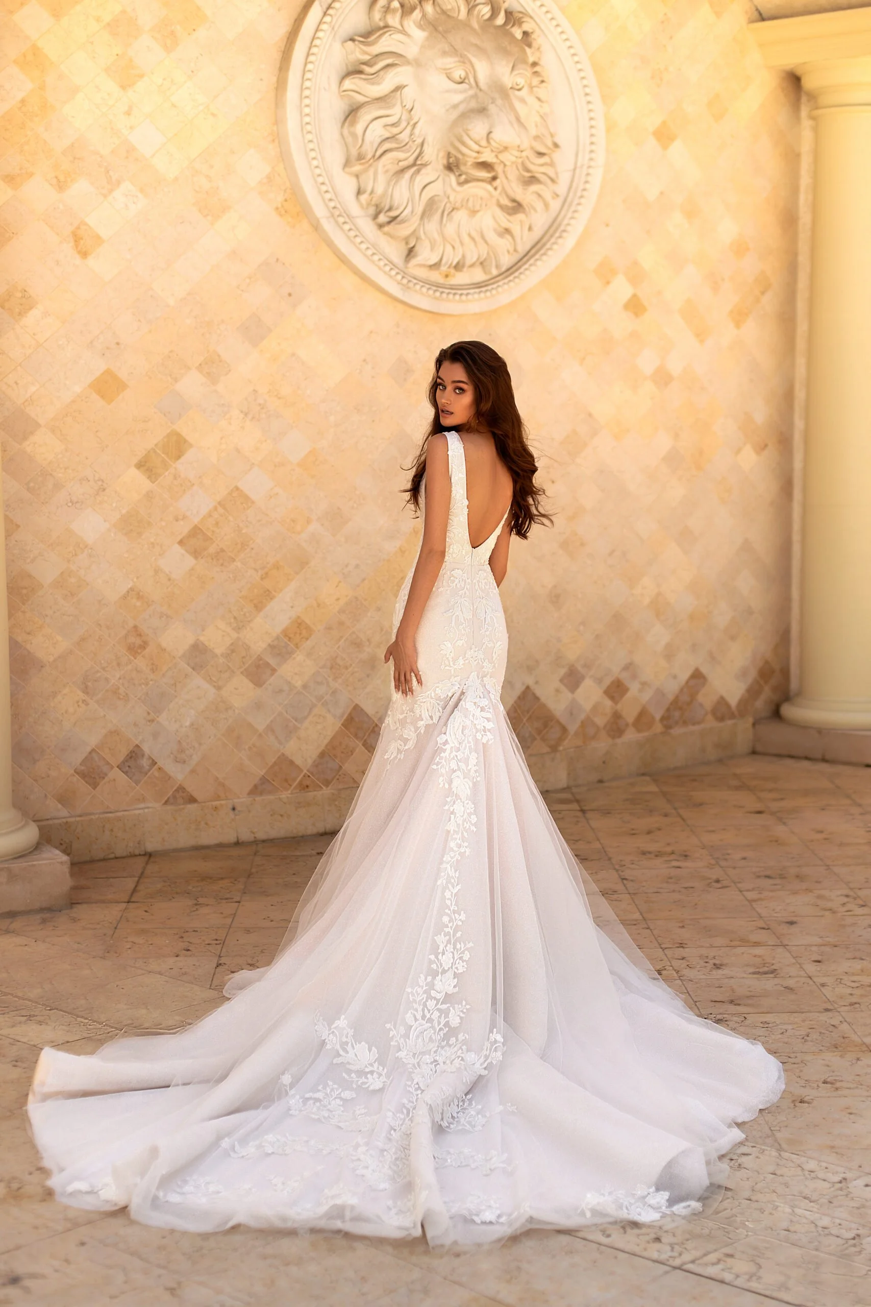 A woman in a white wedding gown with lace details, standing indoors in front of a beige tile wall with a decorative lion face sculpture.