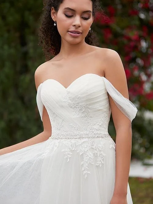 A woman in a strapless white wedding gown with floral lace details, standing outdoors with a blurred green and red floral background.