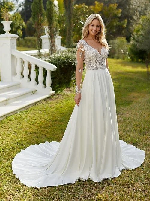 A woman in a white wedding gown with lace long sleeves and a flowing skirt, standing outdoors on grass near a white staircase and railing, with trees and greenery in the background.