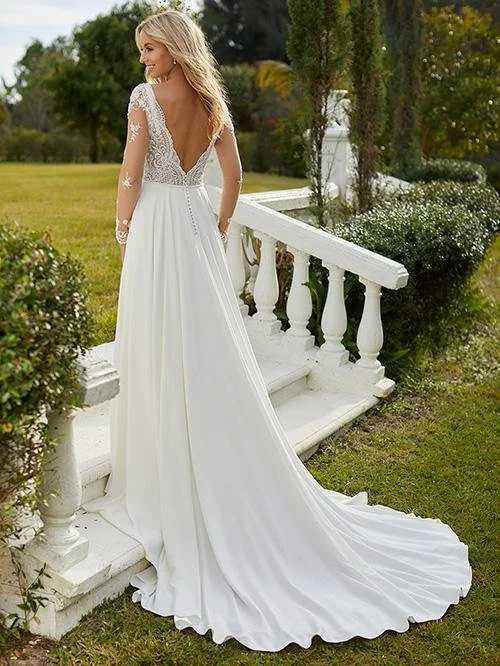 A woman in a white wedding dress with lace sleeves and a long train stands outdoors near a white railing and greenery.