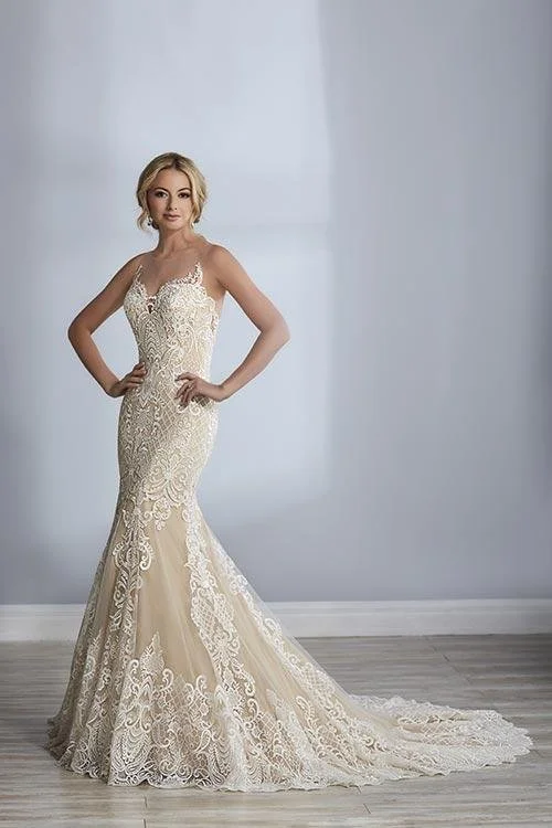 A woman in an elegant, strapless white lace wedding gown with a long train, standing against a plain light gray wall.